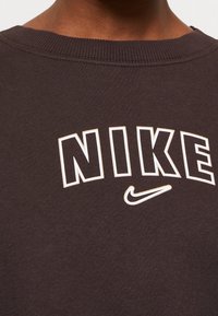Zbliżenie na dekolt czarnej polaru z białym napisem NIKE i małym logo Nike swoosh poniżej liter.