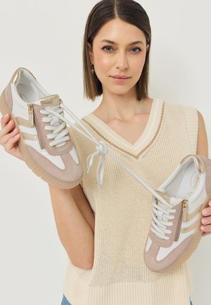 Vrouw met kort bruin haar die een beige gebreid vest draagt, houdt een paar beige en witte sneakers vast met de veters aan elkaar geknoopt.