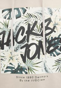 Beige hættebluse med et tropisk blad- og blomsterprint, med "JACK & JONES" tekst i sort. Teksten er fed; designet kombinerer flere farver.