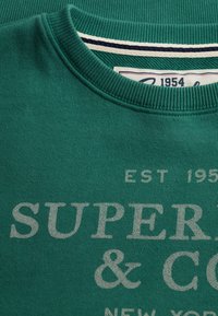 Grøn crewneck-sweatshirt med ribbet krave og falmet hvid tekst, der står "EST 1954 SUPER & CO NEW YORK" på forsiden.