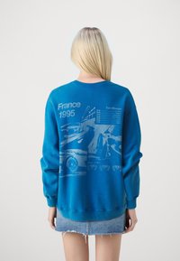 Blauwe sweatshirt met een grafisch ontwerp van auto's en de tekst "Frankrijk 1995" en "Tour d'Europe", met een klassieke pasvorm en geribde mouwen.