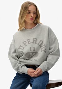 Sudadera gris de gran tamaño con puños acanalados, que presenta "Superdry 1986 Athletic" impreso en letras grandes y contrastantes en la parte frontal.
