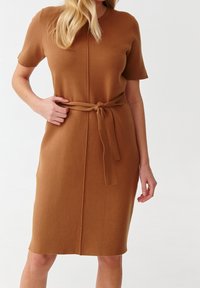 Neulepuku lämpimässä ruskeassa värissä, jossa on lyhyt hihat, istuva vyötärö ja sidottu vyö. Silkkinen tekstuuri, polvipituinen, minimalistinen muotoilu.