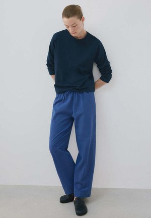 Femme aux cheveux attachés en arrière portant une chemise bleu marine à manches longues, un pantalon bleu à carreaux et des chaussures noires, debout contre un mur blanc uni.