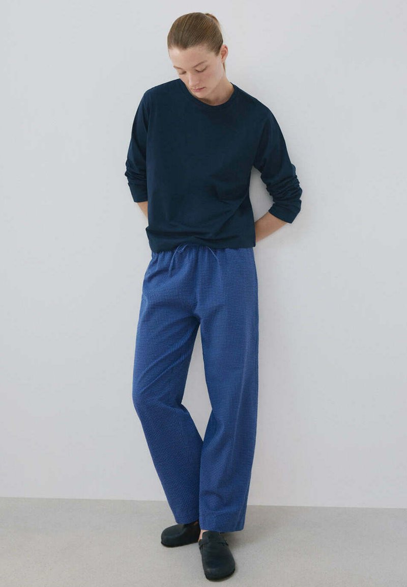 Femme aux cheveux attachés en arrière portant une chemise bleu marine à manches longues, un pantalon bleu à carreaux et des chaussures noires, debout contre un mur blanc uni.