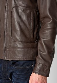 Giubbotto in pelle marrone con una texture liscia, dotato di una tasca laterale e di dettagli in cucitura lungo l'orlo e i polsini.