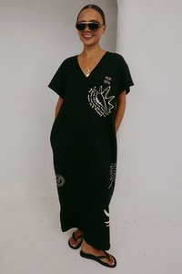 Czarna maxi sukienka z dekoltem w serek i krótkimi rękawami, ozdobiona białymi nadrukami graficznymi muszli i tekstu. Noszona z klapkami i okularami przeciwsłonecznymi.