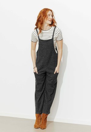Femme aux cheveux roux debout, les mains dans les poches, portant une chemise rayée à manches courtes, une salopette gris foncé et des bottines marron sur fond blanc.