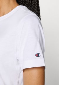 Weißes Baumwoll-T-Shirt mit Rundhalsausschnitt und kurzen Ärmeln. Mit einem kleinen Logo in Rot und Blau auf dem linken Ärmel. Glatte Textur.