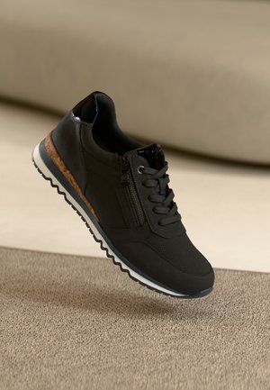 Sneaker nera con lacci e cerniera laterale, suola testurizzata con rifiniture bianche e di sughero, appoggiata su un tappeto beige con sfondo beige sfocato.