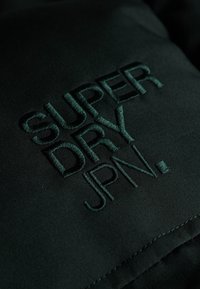 Zwarte stof met donkergroene geborduurde tekst die "SUPER DRY JPN" leest, met een zachte textuur en zichtbare stiksels.
