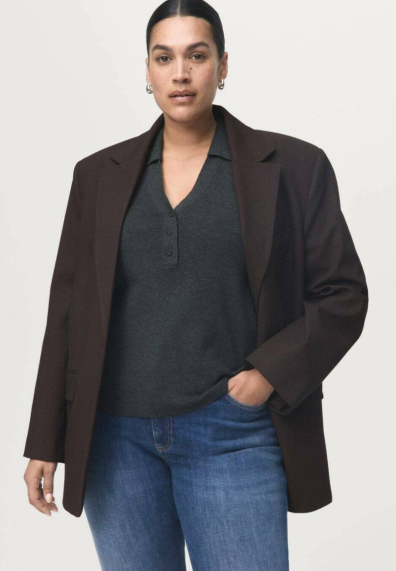 Mujer con cabello oscuro recogido en un moño bajo lleva pendientes dorados, blazer marrón oscuro sobre camisa gris antracita con cuello y vaqueros azules con una mano en el bolsillo.