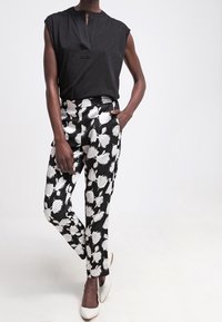 Schwarzes ärmelloses Oberteil mit einem dezenten Kragen, kombiniert mit schwarz-weißen, floral gemusterten Hosen. Weiße Schuhe runden das Outfit ab.