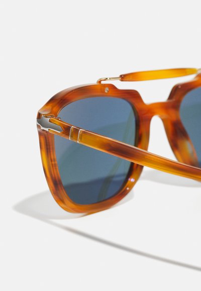 Ochelari de soare cu ramă groasă din scoică, lentile albastre și accesorii aurii. Designul are o formă îndrăzneață și o finisare lucioasă.