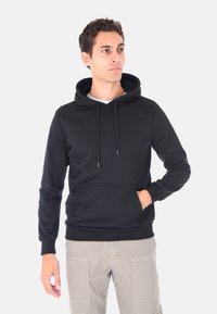 Uomo in felpa nera con tasca frontale e pantaloni cargo beige, in piedi con una mano in tasca, che guarda a destra su uno sfondo bianco.