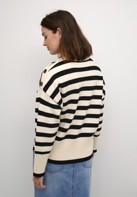 Pull rayé en crème et noir, avec une coupe ample, des poignets côtelés et des accents de boutons sur l'épaule. Texture tricot douce.