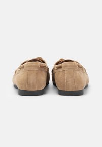 Brune semskede loafers har et glat design, avrundet form og svarte gummisåler med dekorative metallaccent på overdelen.