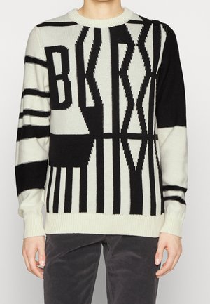 Person trägt schwarzen und weißen Strickpullover mit großen vertikalen Streifen und fetten Blockbuchstaben, die "BKNY" auf der Vorderseite buchstabieren.