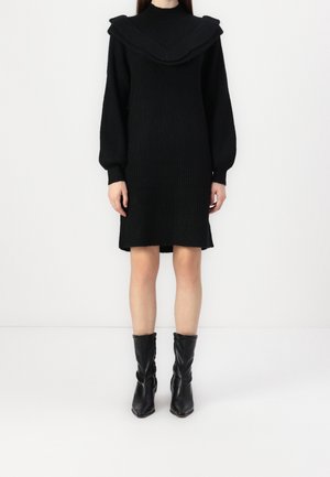 Nainen pukeutuneena mustaan ribbineulottuun mekkoon, jossa on puffihihat ja leveä röyhelökaulus, yhdistetty mustiin nahkasaappaisiin keskisääreen asti.