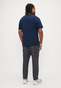 Μπλε σκούρο κοντομάνικο μπλουζάκι με στρογγυλή λαιμόκοψη, σε συνδυασμό με γκρι παντελόνι cargo με πλαϊνές τσέπες. Το μοντέλο φοράει αθλητικά παπούτσια σε ουδέτερους τόνους.