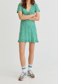 Vestido floral verde con escote en V y mangas cortas, que presenta un dobladillo acampanado. Combinado con zapatillas grises y calcetines blancos con rayas de colores.