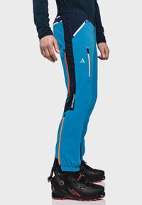 Niebieskie i granatowe spodnie narciarskie z pomarańczowymi zamkami błyskawicznymi, o dopasowanym kroju i wzmocnionych mankietach przy kostkach. Posiadają wiele kieszeni na zamki.