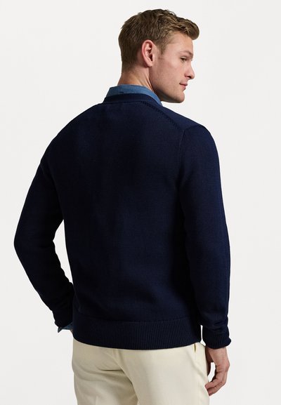 Marinblå stickad tröja med ribbade mudd och nederkant. Har en rund hals och ett texturerat mönster. Bärs över en ljusblå skjorta med krage.