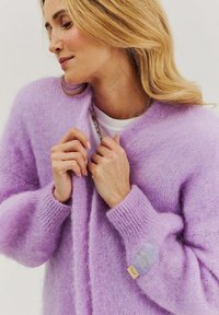 Levanduľový nadýchaný kardigán s dlhými rukávmi. Má štítok na ľavom rukáve a otvorený predný dizajn. Mäkká, textúrovaná látka.