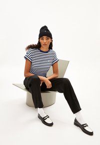 Gestreept T-shirt in marineblauw en wit, met opgerolde mouwen, gecombineerd met zwarte broek, witte sokken en zwarte instappers. Zittend op een lichtgrijze stoel.