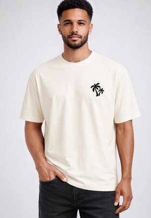 Mann mit kurzen lockigen Haaren und Bart trägt ein cremefarbenes T-Shirt mit einem kleinen schwarzen Palmenmotiv auf der linken Brust, kombiniert mit schwarzen Hosen.