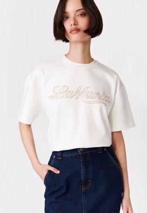 Kobieta o krótkich ciemnych włosach, ubrana w białą koszulkę "La Mania" i ciemnoniebieskie jeansy z wysokim stanem, ręka w kieszeni, na jednolitym tle.