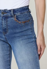 Korkeavyötäröiset siniset farkut, joissa on hienovarainen brodeeraus etutaskussa ja pieniä koristeellisia yksityiskohtia sauman varrella.