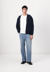 Marineblå zip-up hoodie over en hvid t-shirt. Lyseblå jeans i lige snit med subtil falming. Sorte sko. Ensfarvet baggrund.