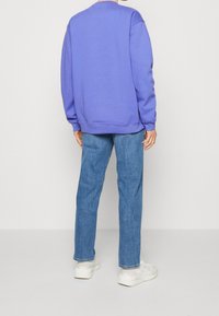 Felpa viola con collo a giro, vestibilità rilassata e polsini a costine, abbinata a jeans in denim blu e sneaker bianche. Mostrata vista posteriore.