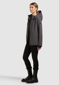 Jeune femme portant une veste grise à capuche, des leggings noirs et des bottes noires épaisses, debout de profil sur un fond uni.