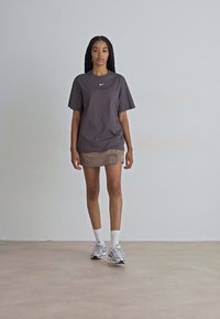 Grå T-skjorte med en hvit Nike-logo, parret med et beige skjørt. Modellen har på seg hvite sokker og grå joggesko. Enkelt bakgrunn.