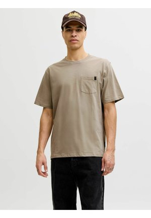 Jeune homme portant un T-shirt beige à poches, un pantalon noir et une casquette marron foncé avec un motif jaune, debout devant un fond clair uni.