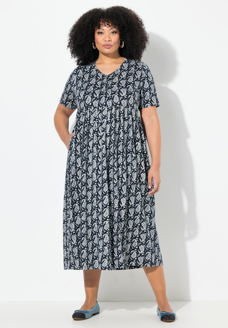Schwarz-weißer Paisley-Muster-Kleid mit kurzen Ärmeln, einem V-Ausschnitt und gerafftem Taillenbund, das bis unter das Knie fällt. Flache blaue Schuhe.