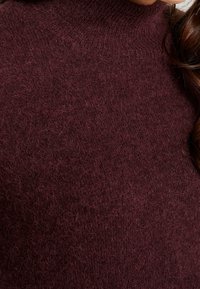 Jersey de punto color granate con un cuello alto, que presenta una textura suave y esponjosa y detalles acanalados en el cuello y el dobladillo.