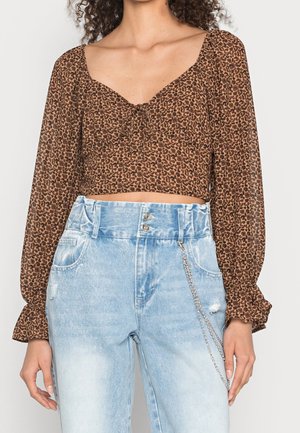 Kvinde iført brun leopardprint crop top med lange ærmer og lyseblå højttaljede jeans med kæde-tilbehør.
