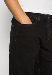 Vaqueros de denim negro con un diseño de pierna recta, costuras en contraste y bolsillos laterales. La textura parece suave con un ligero brillo.
