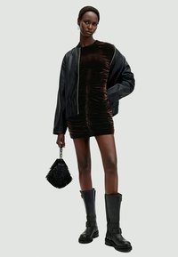 Casaco bomber preto sobre um vestido de veludo castanho com franzido. Botas altas pretas até ao joelho e uma mala preta texturizada com uma pega de corrente.