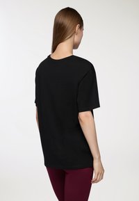 T-shirt nera in cotone con collo rotondo e maniche corte, caratterizzata da una vestibilità ampia. La visuale dal retro mostra un design semplice e una texture liscia.