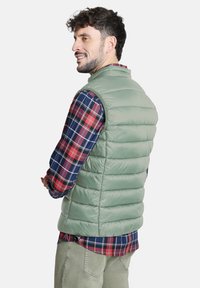 Hombre con chaleco acolchado verde claro sobre una camisa de cuadros roja y azul y pantalones beiges, de perfil con una ligera sonrisa.