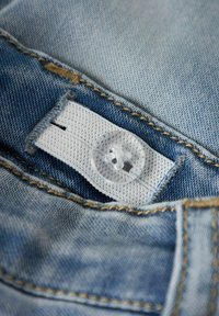 Blå denimstoff med synlige sømmer, med en hvit etikett og en klar plastknapp. Teksturen ser myk og lett slitt ut.