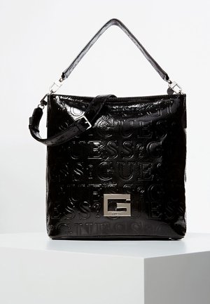 Black patent leather Guess handbag with embossed logo pattern, silver G-shaped clasp, and adjustable shoulder strap on white pedestal.