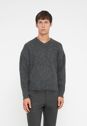 Gray knitted sweater with a V-neckline, dropped shoulders, and a textured finish. Worn over dark gray tailored pants.