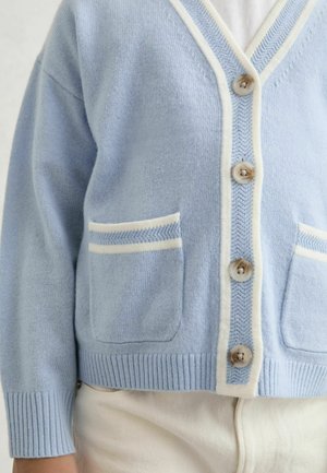 Világoskék kötött kardigán V-nyakkivágással, fehér szegéllyel, négy gombbal és két első zsebbel, amelyek fehér díszítéssel és bordázott mandzsettával rendelkeznek.