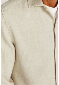 Beige tekstureret jakke med krave og knapper, båret over en hvid ribbet crew neck T-shirt på en person.