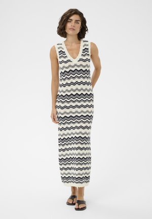 Žena s krátkymi kučeravými vlasmi, oblečená v bezrukávových dlhých pletených šatách s čierno-bielymi vlnitými pruhmi, stojaca so zdvihnutými rukami za chrbtom, čierne sandále.